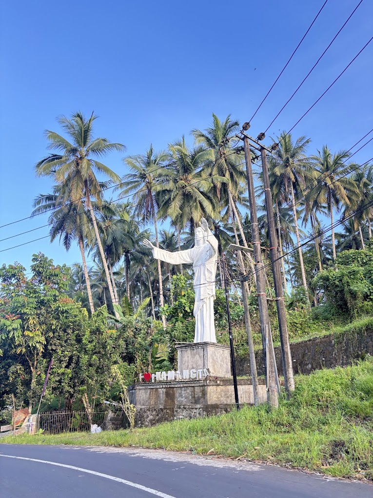 Pemandangan Desa Kamangta dengan pepohonan kelapa dan penanda kawasan di tepi jalan